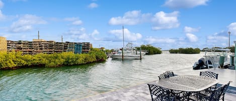 Een terras aan het water met een ronde tafel en stoelen, een steiger met boten, en een gebouw met gestapelde kratten.