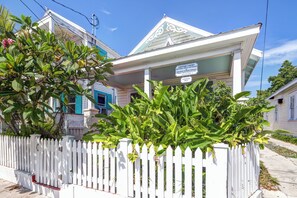 A white house with a sign indicating it is a historical site, surrounded by a white picket fence and lush greenery.
