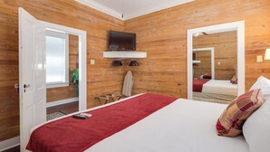 A bedroom with a wooden wall panel, a bed with a red blanket, a TV mounted above the bed, and a mirror reflecting the room.