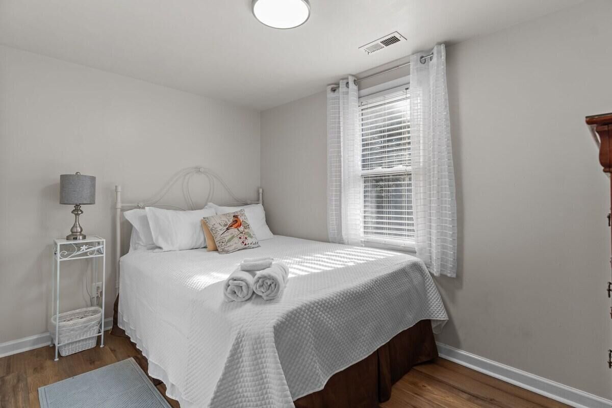 A bedroom with a bed, white curtains, a bedside lamp, and a wooden floor.