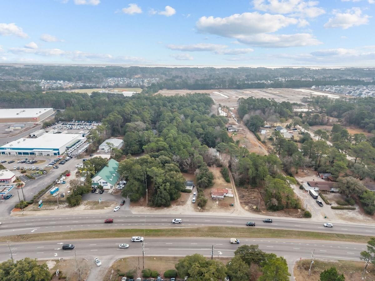 An aerial view of a highway with multiple lanes and vehicles. There are buildings, including a large white structure with a blue roof, and a smaller building with a green roof.