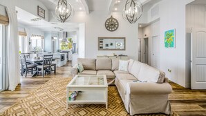 Amplia sala de estar con un sofá color beige, una mesa de centro y un tapete estampado. Se observa un comedor con mesa y sillas, y una cocina al fondo.
