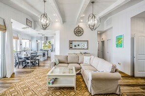 A spacious living room with a beige sofa, a coffee table, and a patterned rug. There is a dining area with a table and chairs, and a kitchen in the background. - Amazing waterfront home with private beach, dock, pool, WiFi & outdoor kitchen (Cudjoe Key)