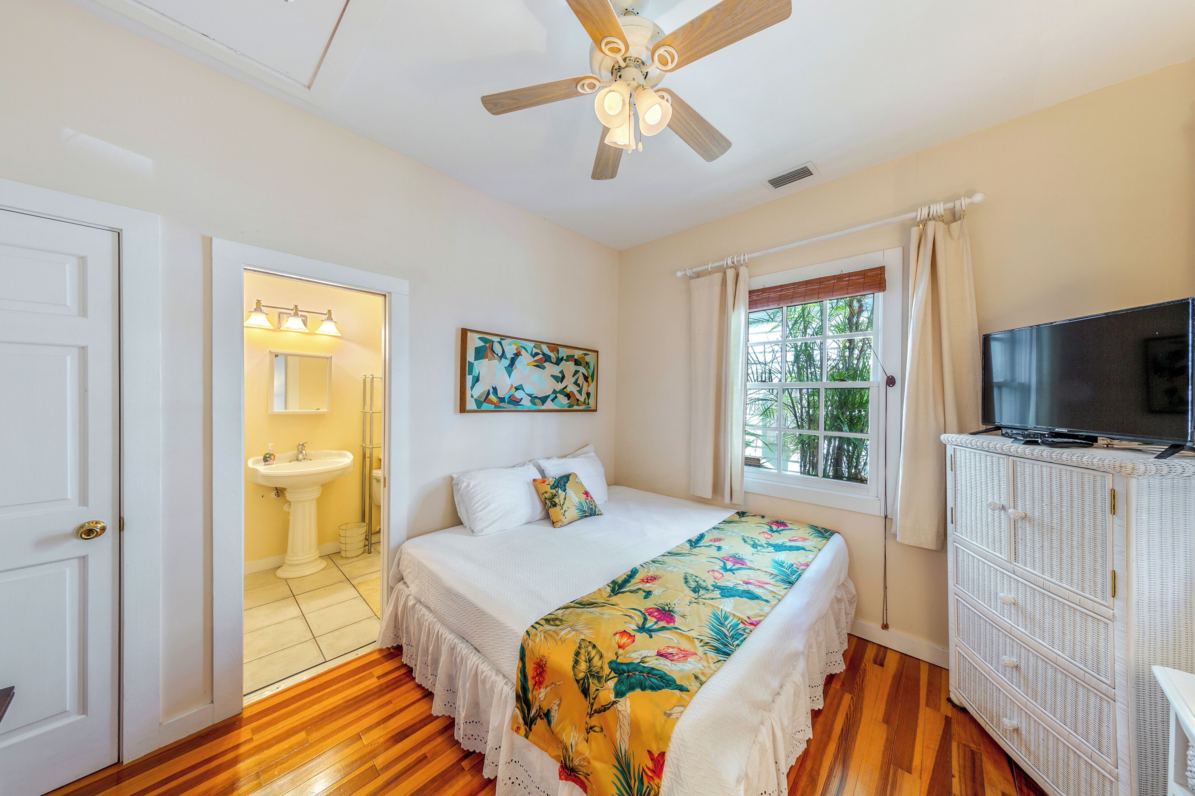 A bedroom with a bed, a ceiling fan, a television, a window with curtains, and a door leading to a bathroom.