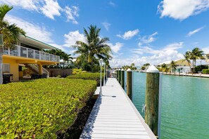 A waterfront walkway with a wooden deck, palm trees, and a building with a balcony. - Waterfront home with dock, filet station, patio, balcony, & Cabana Club access (Key Colony Beach)