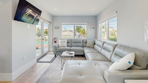 Una sala de estar moderna con un sofá seccional gris, una mesa de centro y un ventanal con vistas a una piscina y follaje.