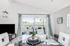 A modern living room with a glass table, white chairs, a TV, and a view of a pool and palm trees. - Enchanting dog-friendly waterfront retreat with pool, hot tub, & amazing views (Key Colony Beach)