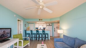A living room with a TV, a white coffee table with a flower, a blue sofa, and a ceiling fan.