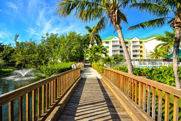 A wooden boardwalk leading to a hotel with a green roof, surrounded by lush greenery and palm trees.