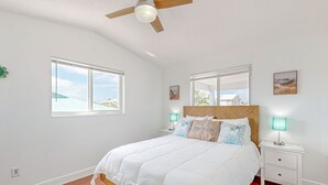 Een slaapkamer met een houten bedframe, wit beddengoed, twee nachtkastjes met lampen, en grote ramen met uitzicht op gebouwen en groen.