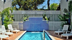 A swimming pool with a tiled wall, surrounded by white lounge chairs and a fence with plants. - Cute Dog-friendly Tropical Condo with AC, WiFi, & Shared Pool - Snowbirds OK (Key West)