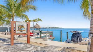 A beachside view with a sign for Ocean Isles Fishing Village, a dock with boats, and palm trees. - Condo in Marathon with waterfront pool, patio, kayak & sundeck (Marathon)