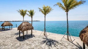 A beachside area with palm trees and thatched umbrellas, a paved walkway, and a view of the sea. - Condo in Marathon with waterfront pool, patio, kayak & sundeck (Marathon)