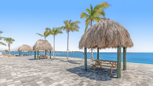 Une zone aménagée au bord de la plage, avec des parasols en chaume et des bancs en bois, une promenade dallée et des palmiers.