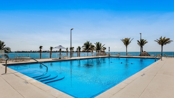 Une piscine rectangulaire à l’eau d’un bleu cristallin, entourée d’une terrasse en béton, de palmiers et de chaises longues.