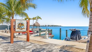 A beachside view with a sign for Ocean Isles Fishing Village, a dock with boats, and palm trees. - Snowbird-friendly sound view condo with open layout, patio, W/D, & AC (Marathon)