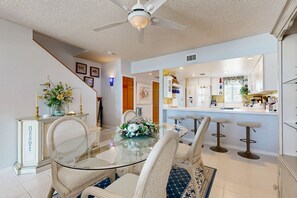 A dining area with a glass table, white upholstered chairs, a ceiling fan, and a kitchen in the background. - Historically located condo w/ocean views, pool, loft, AC & guest boat access (Layton)