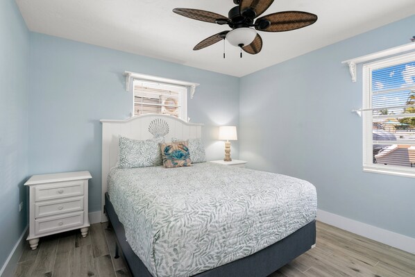 A bedroom with a bed, a nightstand, a ceiling fan, a window with blinds, and a view of the outside.