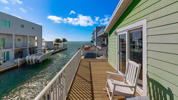 A deck with a view of the ocean, a boat docked on the left, and a building with a green exterior. - Modern waterfront home with balcony, patio, grill, W/D, & easy Atlantic access (Marathon)