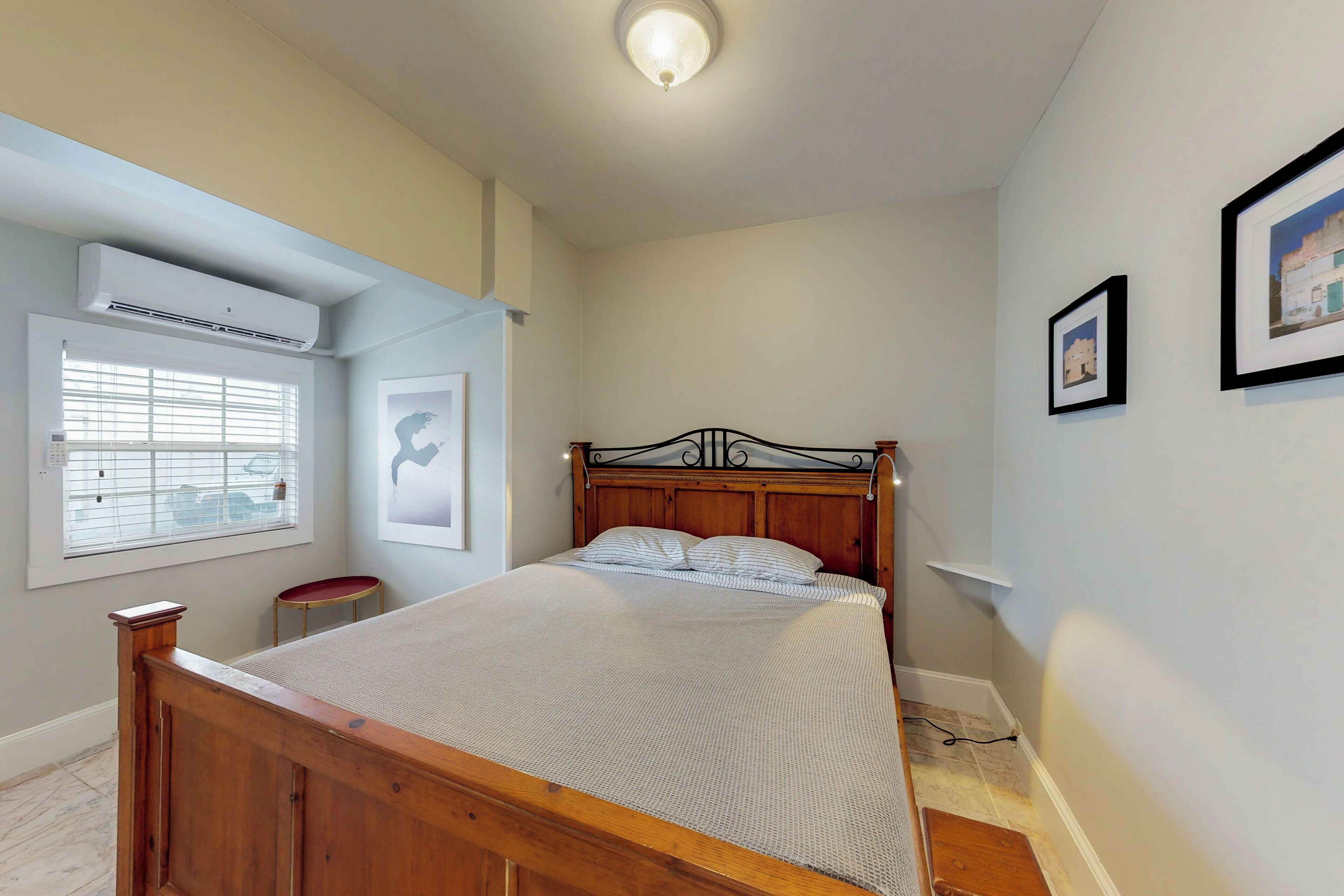 A bedroom with a wooden bed, a window with blinds, a wall-mounted air conditioner, and framed pictures.