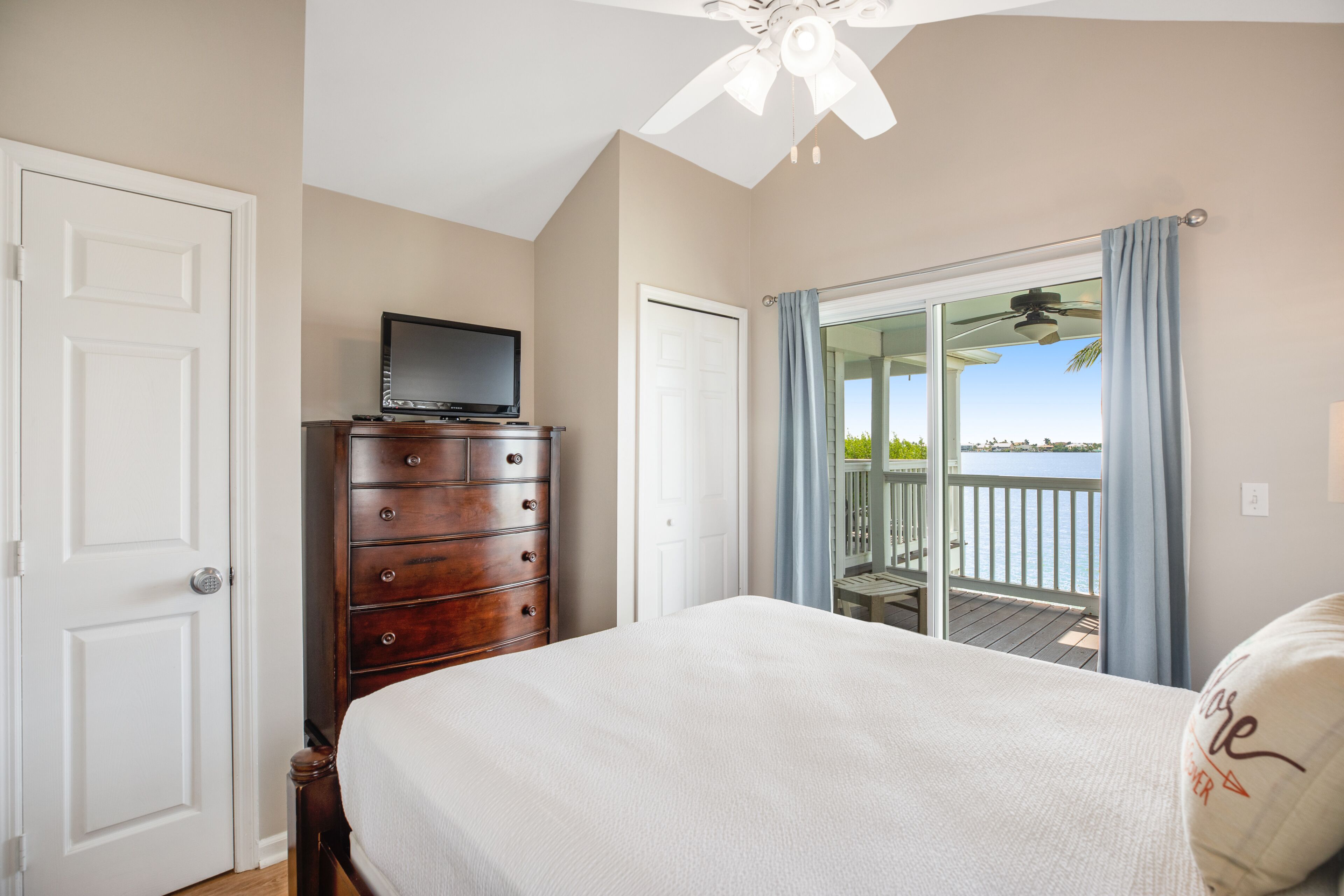 A bedroom with a bed, a dresser, a television on the dresser, and a balcony with a view of the water.