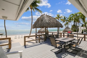 A wooden deck with a table and chairs, thatched umbrellas, and a view of the beach and ocean.