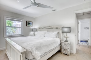 A white bedroom with a bed, a ceiling fan, a window, and a door leading to a bathroom.