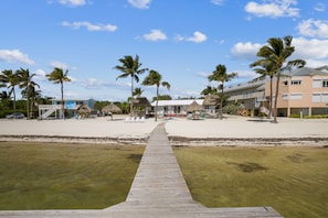 A beachside resort with a wooden walkway leading to a building, palm trees, and a clear sky.