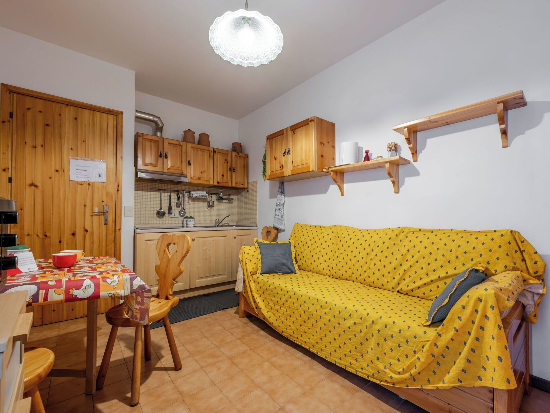 A compact kitchen with wooden cabinets, a dining table with chairs, and a yellow sofa with patterned cushions.