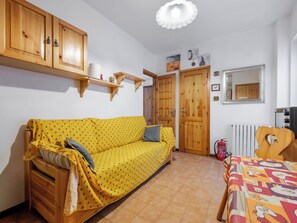 Ein Zimmer mit einem gelb gemusterten Sofa, Holzschränken, einem Heizkörper und einem Bett mit bunter Bettdecke.