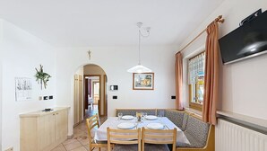 En matplats med ett bord, stolar, en bänk, en tv och ett fönster med gardiner.