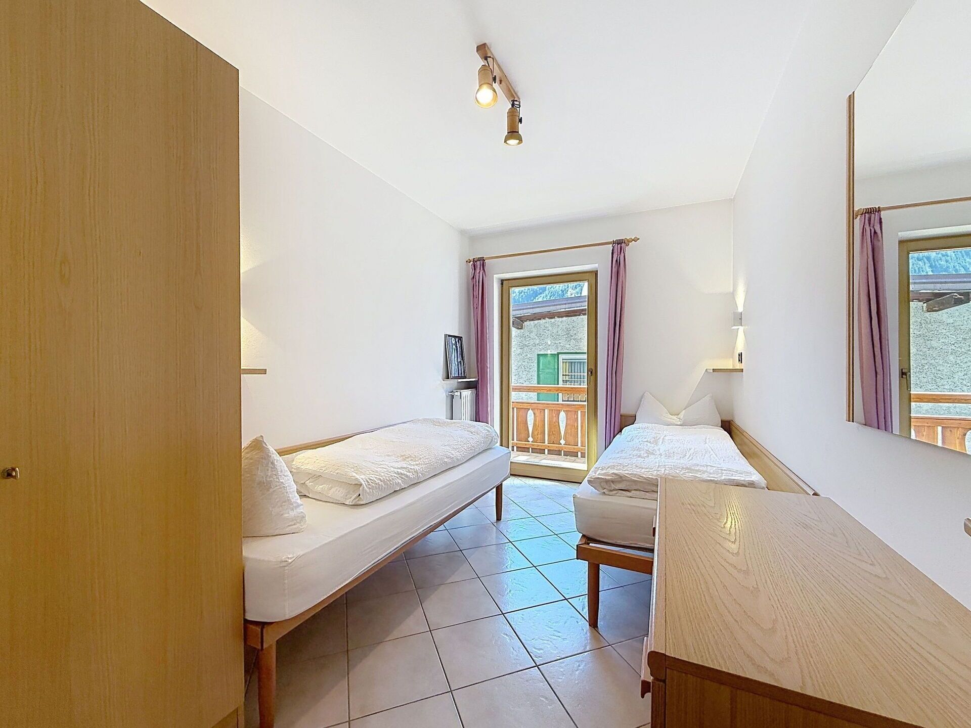 A bedroom with two single beds, a wooden desk, a window with curtains, and a view of a balcony.