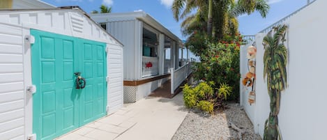 A pathway leading to a house with a turquoise door, a porch, and a small garden with palm trees and shrubs.