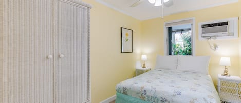 A bedroom with a bed, a wardrobe, a ceiling fan, a window with an air conditioning unit, and a framed picture on the wall.