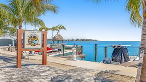 A beachside view with a sign for Ocean Isles Fishing Village, a thatched-roof structure, and boats docked at the pier. - Snowbird-friendly condo in oceanfront complex with heated pool, BBQ, W/D, AC (Marathon)