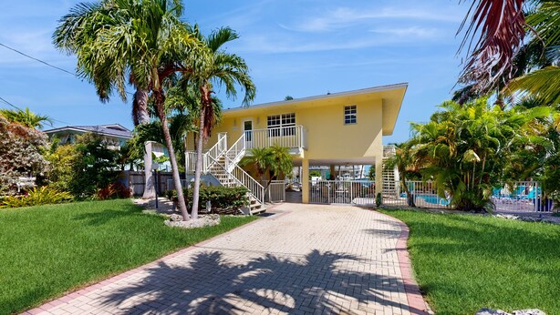 A yellow two-story house with a paved driveway, palm trees, and a fenced pool area. - Private pool, boat dock, water view, canal front, fully equipped kitchen (Marathon)