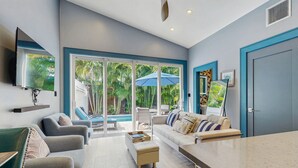 Una sala de estar moderna con un amplio ventanal corredera que da a una zona de piscina, un sofá, una mesa de centro y un televisor montado en la pared.