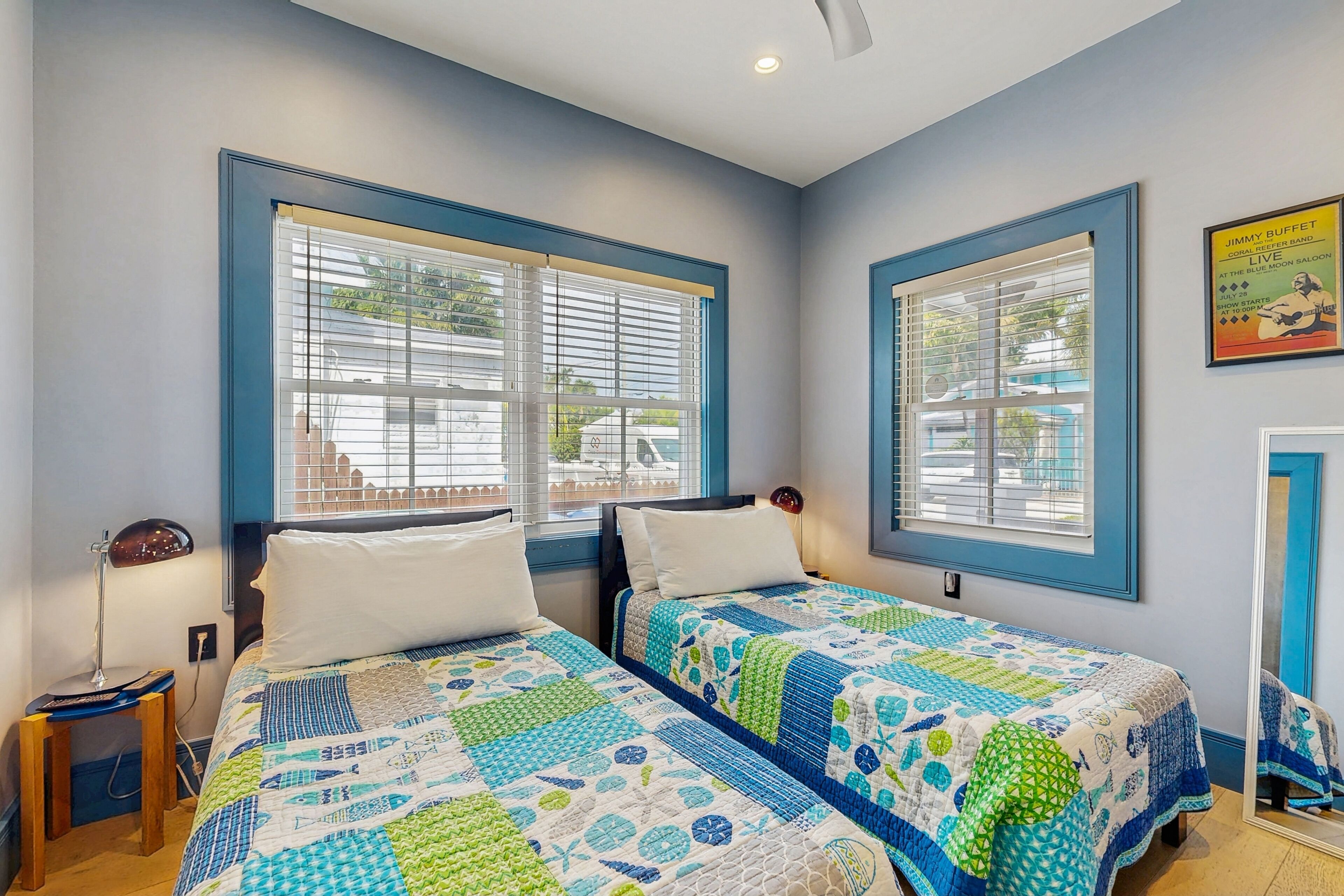 Un dormitorio con dos camas, cada una con colcha azul y blanca, una mesita de noche con lámpara, y un cartel en la pared.