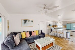 Ein modernes Wohnzimmer mit einem grauen Ecksofa, einem Glastisch und einer Küche im Hintergrund.