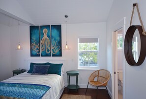 A bedroom with a bed, two wall-mounted paintings, a window, a wicker chair, and a mirror on the wall. - Fresh Getaway w/ Private Pool & Rooftop Patio - Great Location, Steps from Beach (Key West)