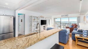 Una cucina moderna con piano di lavoro in granito, frigorifero in acciaio inox e TV a incasso. È presente una zona salotto con divani blu e vista sull'oceano.