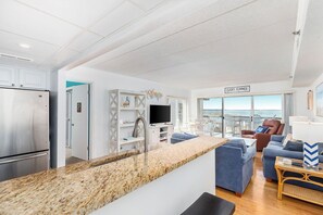 A modern kitchen with a granite countertop, a stainless steel refrigerator, and a built-in TV. There is a seating area with blue sofas and a view of the ocean. - Lovely 3 Bedroom with great bay views, outdoor pool, and just steps to the beach. Vacation at the Shore with Shoreline Properties (Ocean City)