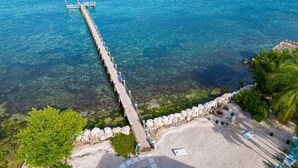 Un muelle se adentra en aguas azules y transparentes, rodeado de una playa arenosa con algunos árboles y un muro de piedra.