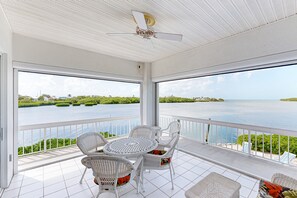 A covered balcony with a table and chairs, overlooking a body of water with greenery and distant buildings. - Luxurious waterfront home with panoramic views, pool, spa, & dock (Marathon)