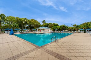 A rectangular swimming pool with a tiled deck, surrounded by lounge chairs and umbrellas, with palm trees in the background. - Breezy condo on the water with pool, hot tub & tennis court (Tavernier)