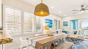 A spacious living room with a large wooden dining table, white chairs, a sofa with blue cushions, and a view of the ocean through large windows. - Waterfront, dog-friendly home with beautiful canal view & 2 shaded porches (Duck Key)