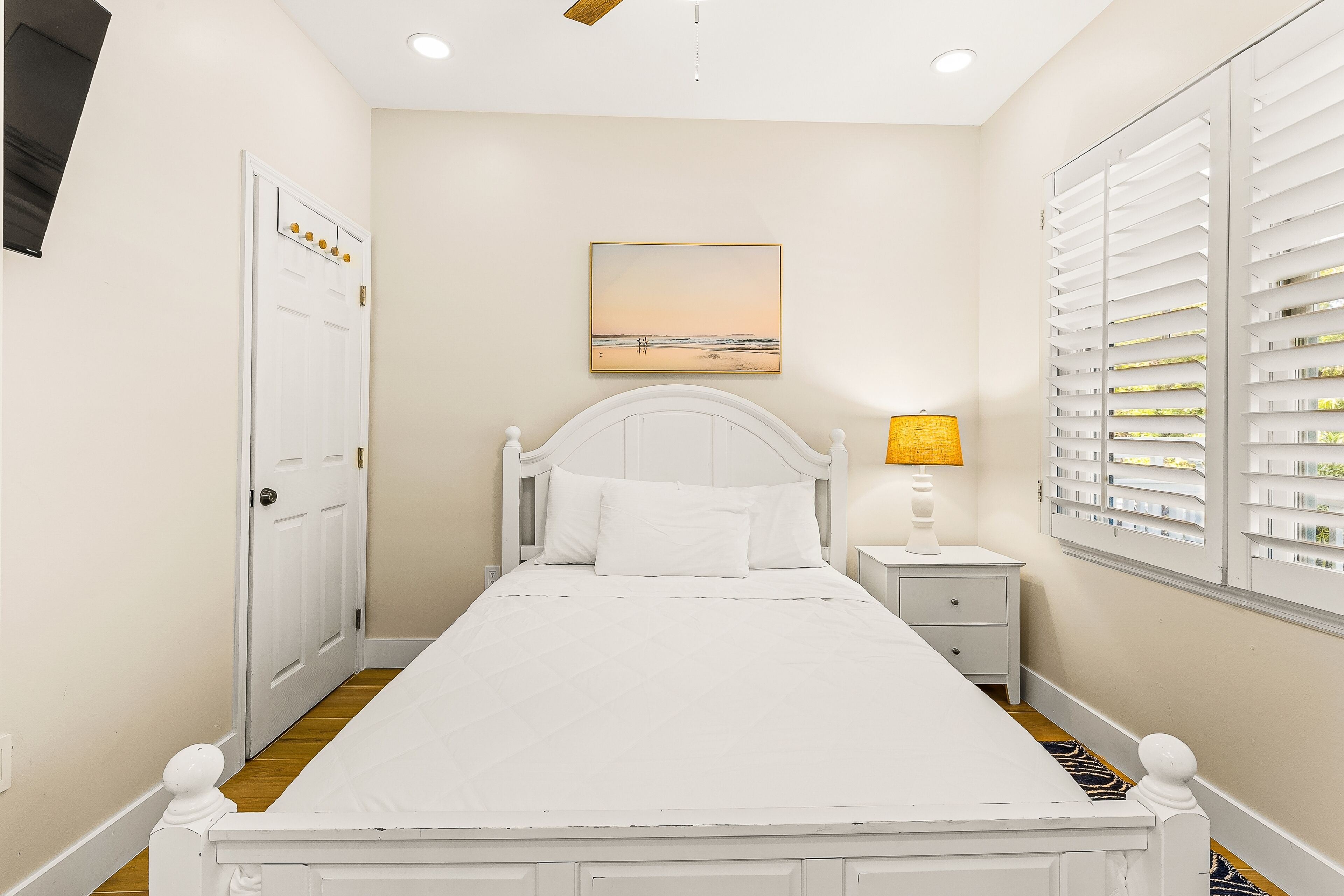 A bedroom with a bed, a nightstand, a lamp, a painting, and a window with blinds.