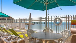 En terrasse ved stranden med hvide liggestole, et rundt bord og en grøn parasol.