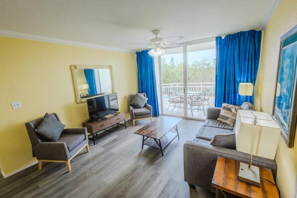 A living room with a sofa, armchairs, a coffee table, a TV, and a balcony with blue curtains. - Resort suite with pool, hot tub, tennis & full kitchen (Key West)