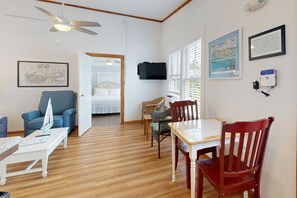 A room with a wooden floor, a dining table with chairs, a blue armchair, a bed with a white headboard, a ceiling fan, and a mounted television. - Bright Condo with Homey Feel, Entertainment & Private Parking - Beach Nearby (Key West)
